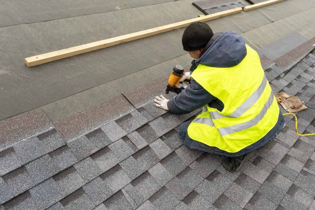 Qualified workman in uniform work wear using air or pneumatic nail gun and installing asphalt or bitumen shingle on top of the new roof under construction residential building