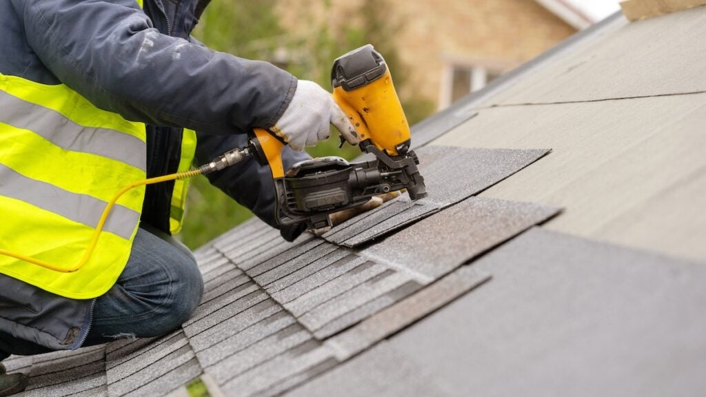 Unrecognizable roofer worker in special protective work wear and gloves, using air or pneumatic nail gun and installing asphalt shingles